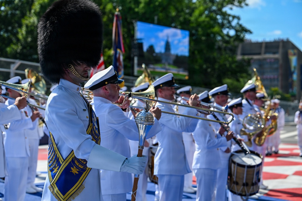 A military marching band performs outdoors in white dress uniforms, with a drum major in the foreground wearing a distinctive tall black bearskin hat and carrying a ceremonial mace. The musicians are playing brass instruments including trombones, French horns, and percussion. The setting appears to be a formal outdoor ceremony or parade on a sunny day.