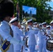 A military marching band performs outdoors in white dress uniforms, with a drum major in the foreground wearing a distinctive tall black bearskin hat and carrying a ceremonial mace. The musicians are playing brass instruments including trombones, French horns, and percussion. The setting appears to be a formal outdoor ceremony or parade on a sunny day.