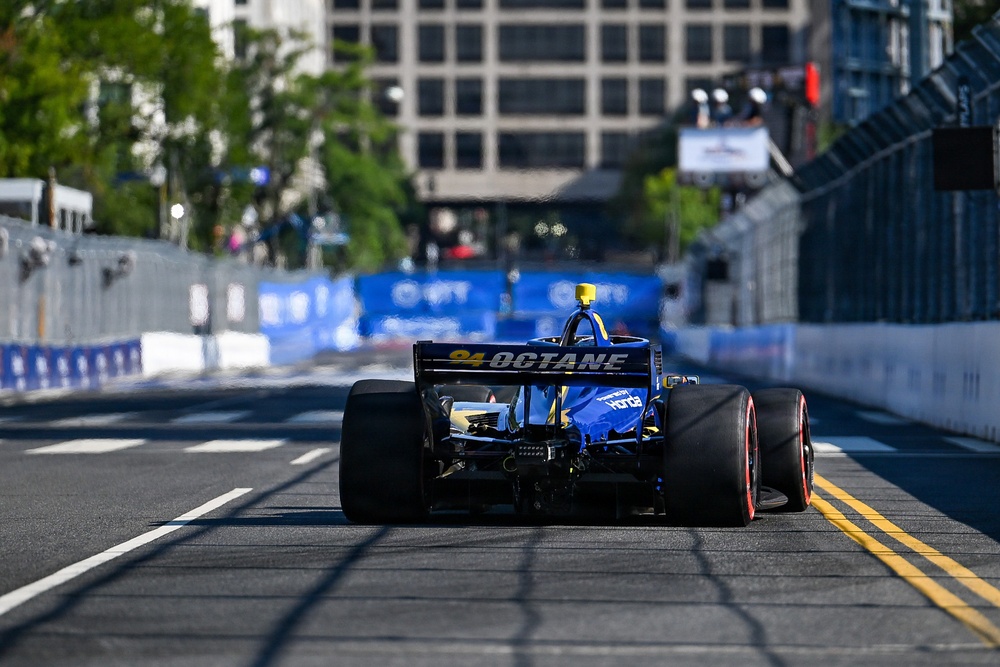 A blue IndyCar racing vehicle is photographed from the rear as it travels down a temporary street circuit lined with concrete barriers. The car displays '94 OCTANE' branding on its rear wing and features yellow accents with red-rimmed rear tires. The urban backdrop includes buildings and trees, consistent with a downtown street race venue.