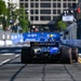 A blue IndyCar racing vehicle is photographed from the rear as it travels down a temporary street circuit lined with concrete barriers. The car displays '94 OCTANE' branding on its rear wing and features yellow accents with red-rimmed rear tires. The urban backdrop includes buildings and trees, consistent with a downtown street race venue.