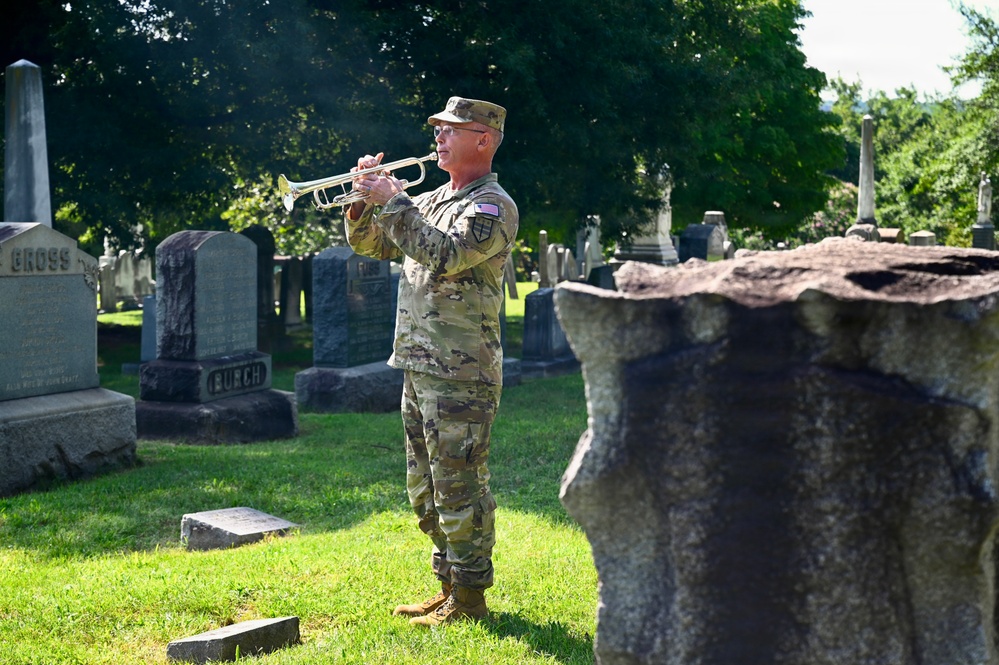 A U.S. Army soldier in OCP (Operational Camouflage Pattern) uniform is playing a bugle or trumpet in a cemetery, likely performing 'Taps' during a memorial ceremony. The soldier stands among numerous headstones on well-maintained grass, with large trees in the background. The scene suggests a military funeral honors or memorial day observance.