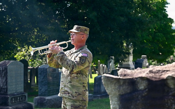 A U.S. Army soldier in OCP (Operational Camouflage Pattern) uniform is playing a bugle or trumpet in a cemetery, likely performing 'Taps' during a memorial ceremony. The soldier stands among numerous headstones on well-maintained grass, with large trees in the background. The scene suggests a military funeral honors or memorial day observance.