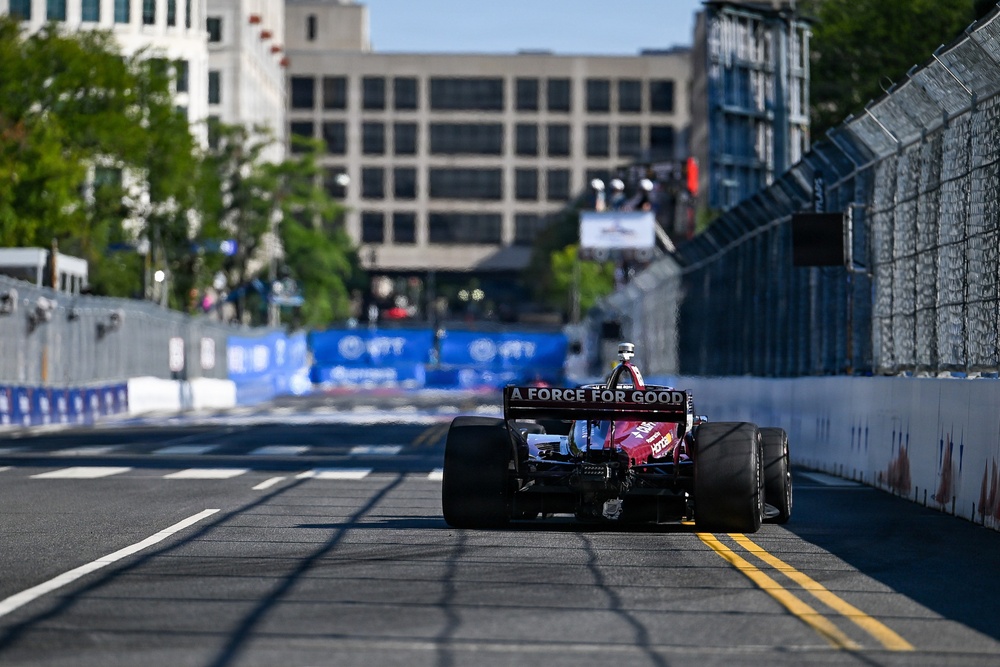 An IndyCar racing car is photographed from behind as it travels down a temporary street circuit lined with concrete barriers and catch fencing. The car displays 'A FORCE FOR GOOD' on its rear wing endplate. The urban backdrop includes multi-story buildings and trees, consistent with a downtown street circuit venue.
