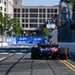 An IndyCar racing car is photographed from behind as it travels down a temporary street circuit lined with concrete barriers and catch fencing. The car displays 'A FORCE FOR GOOD' on its rear wing endplate. The urban backdrop includes multi-story buildings and trees, consistent with a downtown street circuit venue.