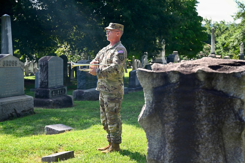 A U.S. Army soldier in MultiCam (OCP) camouflage uniform stands in a cemetery holding a bugle or trumpet, likely preparing to play 'Taps' at a military memorial ceremony. The setting features numerous gravestones and headstones surrounded by lush green trees and grass. This appears to be a formal military honors ceremony, possibly for Memorial Day, Veterans Day, or a funeral service.