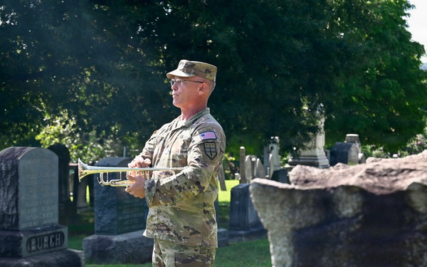 A U.S. Army soldier in MultiCam (OCP) camouflage uniform stands in a cemetery holding a bugle or trumpet, likely preparing to play 'Taps' at a military memorial ceremony. The setting features numerous gravestones and headstones surrounded by lush green trees and grass. This appears to be a formal military honors ceremony, possibly for Memorial Day, Veterans Day, or a funeral service.