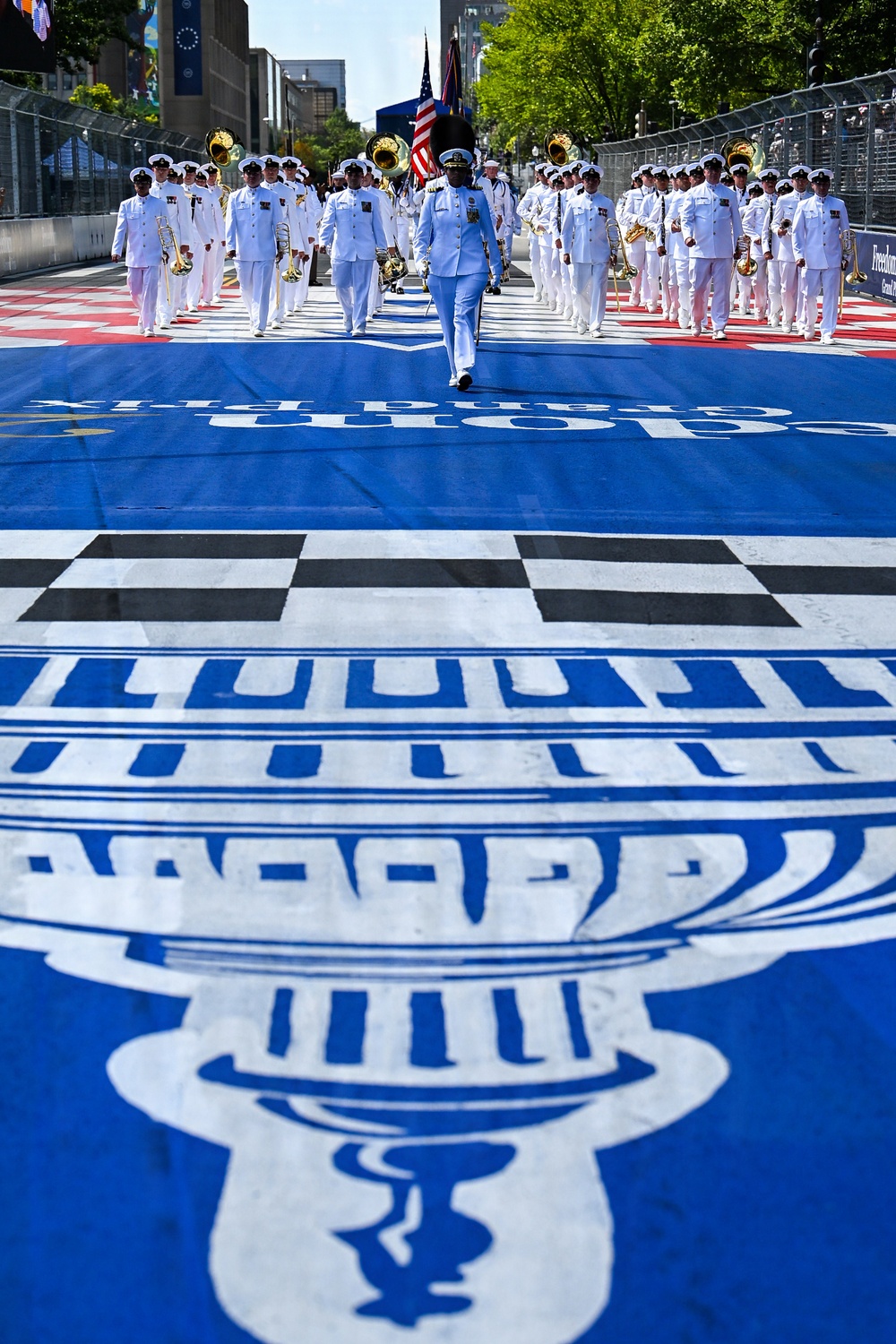 A military marching band in white dress uniforms parades down a racing circuit start/finish straight painted in blue with a large trophy logo and checkered pattern. The procession is led by a drum major and includes brass instrument players carrying an American flag. The setting appears to be a street circuit Grand Prix event, likely the IndyCar Freedom 100 or similar race in an urban environment.