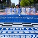 A military marching band in white dress uniforms parades down a racing circuit start/finish straight painted in blue with a large trophy logo and checkered pattern. The procession is led by a drum major and includes brass instrument players carrying an American flag. The setting appears to be a street circuit Grand Prix event, likely the IndyCar Freedom 100 or similar race in an urban environment.