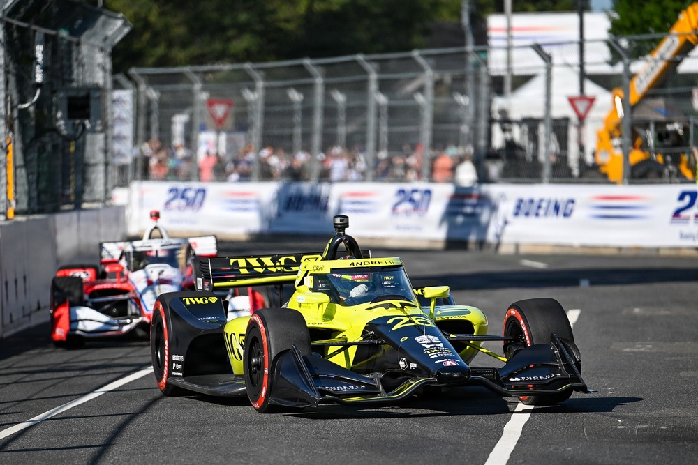 An IndyCar race is underway on a street circuit, with a yellow-and-black Andretti Autosport car bearing the number 26 and TWG sponsorship prominently in the foreground. A red-and-white car follows closely behind. Spectators, safety fencing, and Boeing sponsorship banners are visible in the background.