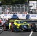 An IndyCar race is underway on a street circuit, with a yellow-and-black Andretti Autosport car bearing the number 26 and TWG sponsorship prominently in the foreground. A red-and-white car follows closely behind. Spectators, safety fencing, and Boeing sponsorship banners are visible in the background.