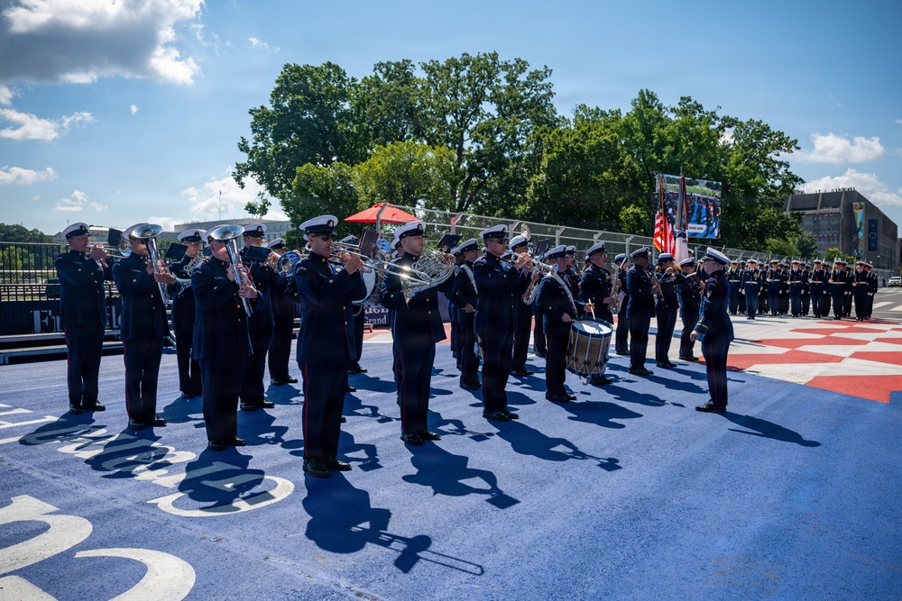 A military band in dark dress uniforms performs outdoors on a decorated blue and red surface, likely at a formal ceremonial event. The musicians are playing brass instruments, French horns, and drums, while a color guard with flags stands to the right. A formation of uniformed personnel is visible in the background.