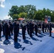 A military band in dark dress uniforms performs outdoors on a decorated blue and red surface, likely at a formal ceremonial event. The musicians are playing brass instruments, French horns, and drums, while a color guard with flags stands to the right. A formation of uniformed personnel is visible in the background.