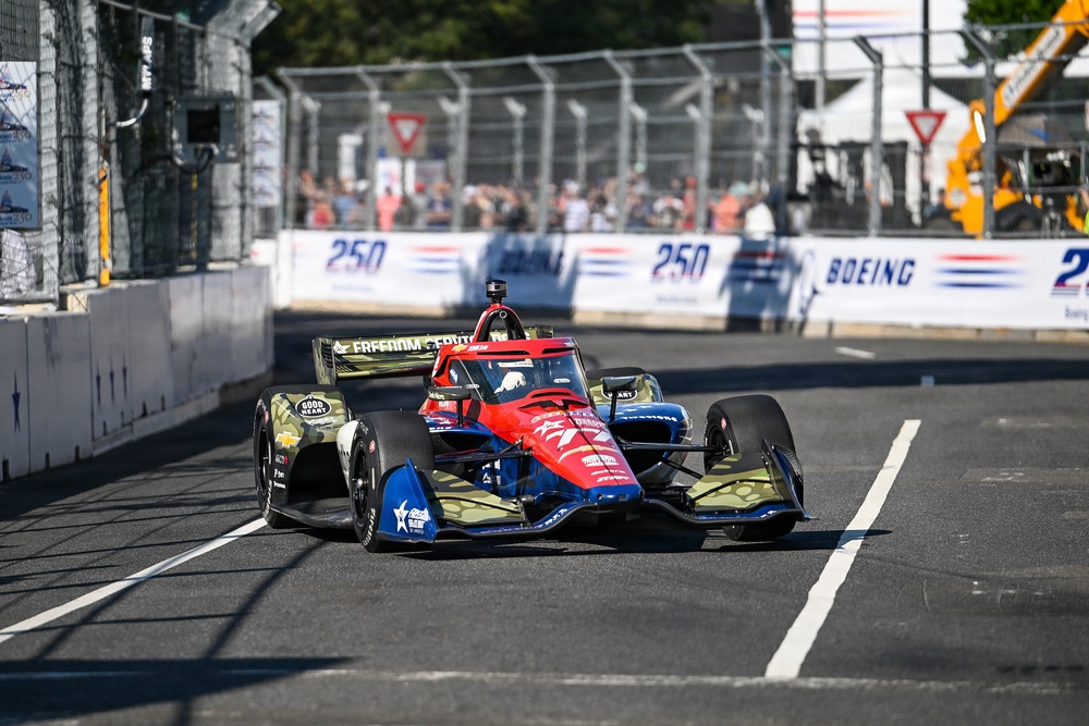 An IndyCar racing car with a red, blue, and military camouflage livery featuring 'Freedom' branding is navigating a street circuit. The car appears to be on track during a race or practice session, with spectators visible in the background behind safety barriers. Sponsor signage including 'Boeing' and '250' is visible on the pit wall.