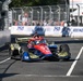 An IndyCar racing car with a red, blue, and military camouflage livery featuring 'Freedom' branding is navigating a street circuit. The car appears to be on track during a race or practice session, with spectators visible in the background behind safety barriers. Sponsor signage including 'Boeing' and '250' is visible on the pit wall.