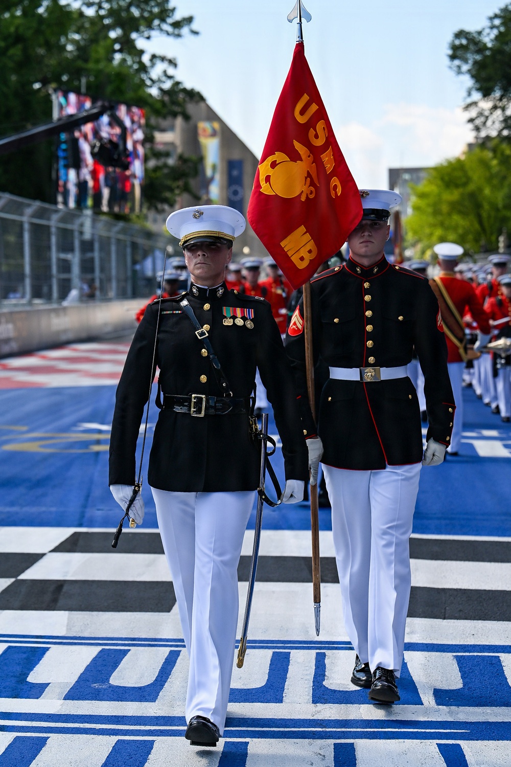 United States Marine Corps (USMC) personnel in dress uniform are marching in a formal parade or ceremonial event on what appears to be a racing circuit or specially marked street course. The lead figure carries a ceremonial sword while a second Marine bears a red USMC guidon flag. Behind them, members of the USMC Marching Band (MB) in red uniforms follow in formation.