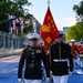 United States Marine Corps (USMC) personnel in dress uniform are marching in a formal parade or ceremonial event on what appears to be a racing circuit or specially marked street course. The lead figure carries a ceremonial sword while a second Marine bears a red USMC guidon flag. Behind them, members of the USMC Marching Band (MB) in red uniforms follow in formation.