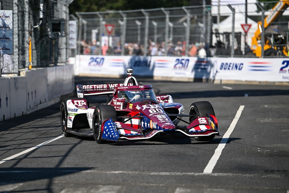 An IndyCar racing vehicle with a patriotic American flag livery competes on a street circuit. The car prominently displays Texas A&amp;M (ATM) and SiriusXM sponsorships along with the phrase 'Together We Stan' on the rear wing. Spectators and safety barriers are visible in the background, along with Boeing and '250' event signage.