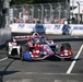 An IndyCar racing vehicle with a patriotic American flag livery competes on a street circuit. The car prominently displays Texas A&amp;M (ATM) and SiriusXM sponsorships along with the phrase 'Together We Stan' on the rear wing. Spectators and safety barriers are visible in the background, along with Boeing and '250' event signage.