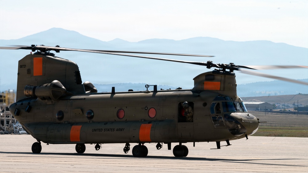 A United States Army CH-47 Chinook tandem-rotor helicopter is parked on a tarmac with its rotors spinning, indicating it is either preparing for takeoff or has just landed. The aircraft bears the tail number 08721 and is painted in olive drab with distinctive orange markings. A mountainous landscape is visible in the background under a hazy sky.