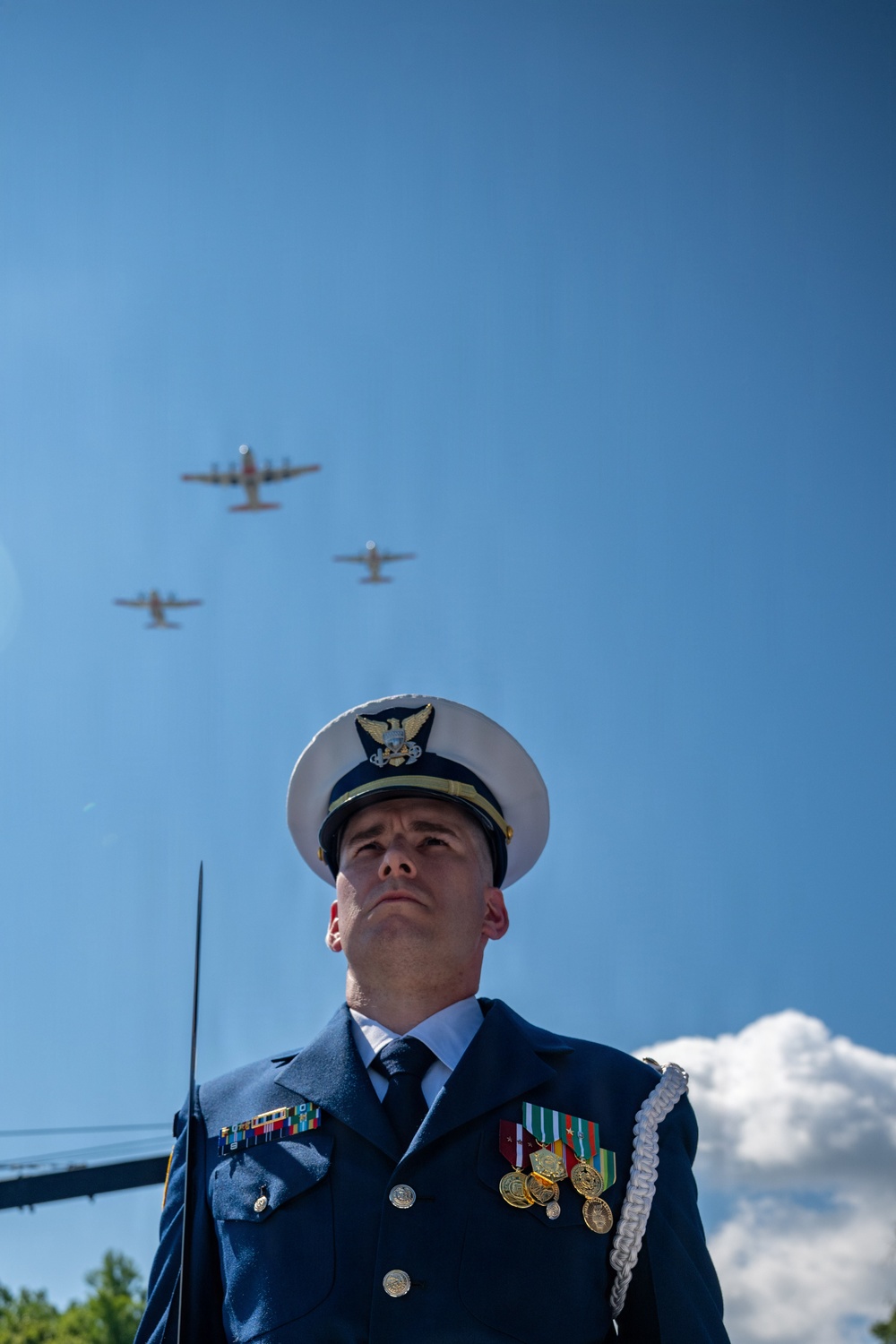 A U.S. Coast Guard officer in dress uniform stands at attention during a formal ceremony while multiple military aircraft conduct a flyover above. The officer is adorned with medals, ribbons, and a ceremonial aiguillette, gazing upward toward the aircraft. The low-angle shot dramatically frames the uniformed figure against a clear blue sky with the formation of planes overhead.