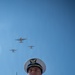 A U.S. Coast Guard officer in dress uniform stands at attention during a formal ceremony while multiple military aircraft conduct a flyover above. The officer is adorned with medals, ribbons, and a ceremonial aiguillette, gazing upward toward the aircraft. The low-angle shot dramatically frames the uniformed figure against a clear blue sky with the formation of planes overhead.