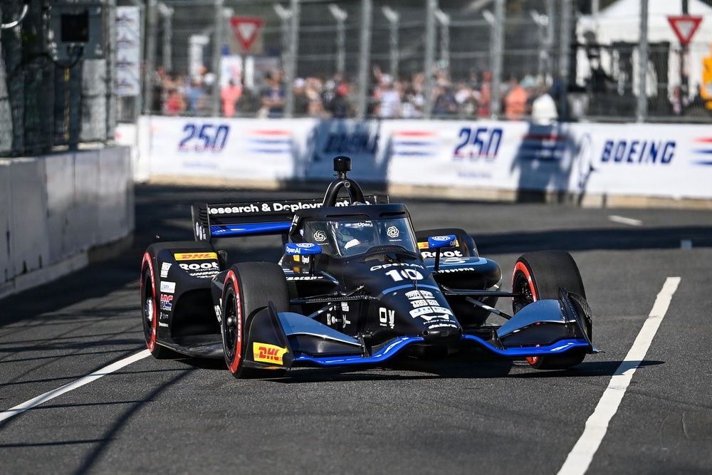 An IndyCar racing car numbered 10 with black and blue livery is navigating a street circuit during a race or practice session. The car features sponsorships including OpenAI, Root, DHL, and 'Research &amp; Deployment' branding. Spectators and barrier walls with '250' and 'Boeing' signage are visible in the background.