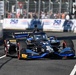 An IndyCar racing car numbered 10 with black and blue livery is navigating a street circuit during a race or practice session. The car features sponsorships including OpenAI, Root, DHL, and 'Research &amp; Deployment' branding. Spectators and barrier walls with '250' and 'Boeing' signage are visible in the background.