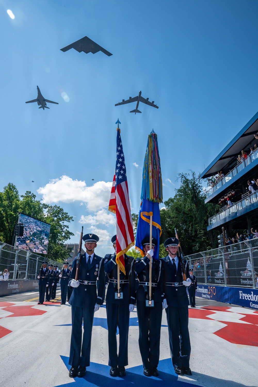 A military color guard ceremony is taking place at the 'Freedom 250 Grand Prix' event, with uniformed Air Force personnel presenting the American flag and a military unit flag on a decorated track surface. Three military aircraft — identifiable as a B-2 Spirit stealth bomber, a B-1B Lancer, and a B-52 Stratofortress — are conducting a flyover directly above the ceremony. Spectators watch from a multi-level grandstand structure on the right side of the image.