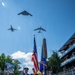 A military color guard ceremony is taking place at the 'Freedom 250 Grand Prix' event, with uniformed Air Force personnel presenting the American flag and a military unit flag on a decorated track surface. Three military aircraft — identifiable as a B-2 Spirit stealth bomber, a B-1B Lancer, and a B-52 Stratofortress — are conducting a flyover directly above the ceremony. Spectators watch from a multi-level grandstand structure on the right side of the image.