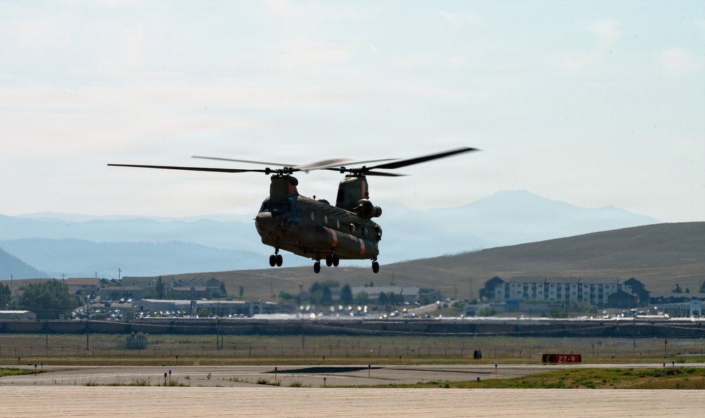 A CH-47 Chinook tandem-rotor military helicopter is captured in low hover or takeoff/landing phase above a paved airfield runway. The aircraft displays olive drab camouflage with orange markings, consistent with U.S. Army livery. A mountainous landscape and small urban area are visible in the background.