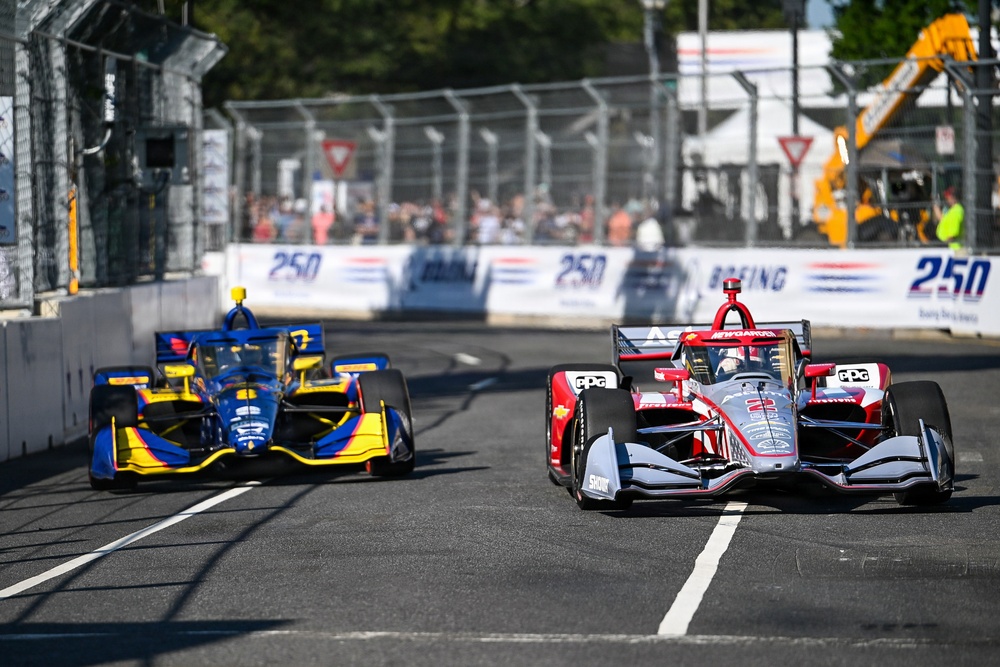 Two IndyCar open-wheel racing cars compete on a street circuit, with a red/silver car leading a blue/yellow car through a straightaway. Sponsor banners including 'Boeing' and '250' are visible along the barrier walls, suggesting this is a sanctioned IndyCar Series event. Spectators, safety fencing, and construction equipment are visible in the background.