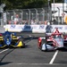 Two IndyCar open-wheel racing cars compete on a street circuit, with a red/silver car leading a blue/yellow car through a straightaway. Sponsor banners including 'Boeing' and '250' are visible along the barrier walls, suggesting this is a sanctioned IndyCar Series event. Spectators, safety fencing, and construction equipment are visible in the background.