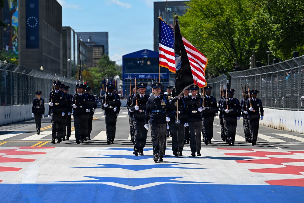 A uniformed honor guard unit marches in formation down a decorated urban street, carrying the American flag and a POW/MIA flag. The procession appears to be part of a formal ceremonial parade, likely in Washington D.C., based on the urban architecture and decorated road surface. The street features a painted red, white, and blue star design, and temporary race barriers line both sides, suggesting a multi-event venue.