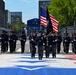 A uniformed honor guard unit marches in formation down a decorated urban street, carrying the American flag and a POW/MIA flag. The procession appears to be part of a formal ceremonial parade, likely in Washington D.C., based on the urban architecture and decorated road surface. The street features a painted red, white, and blue star design, and temporary race barriers line both sides, suggesting a multi-event venue.