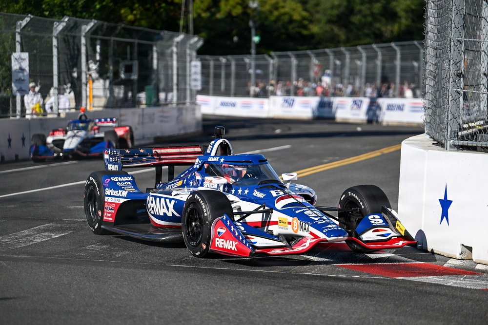An IndyCar race is underway on a street circuit, with the lead car prominently displaying a patriotic American-themed livery sponsored by RE/MAX. The car is navigating a tight corner bordered by concrete barriers and metal fencing. Additional race cars and spectators are visible in the background.