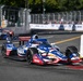 An IndyCar race is underway on a street circuit, with the lead car prominently displaying a patriotic American-themed livery sponsored by RE/MAX. The car is navigating a tight corner bordered by concrete barriers and metal fencing. Additional race cars and spectators are visible in the background.