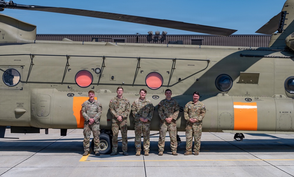 Five military personnel in camouflage uniforms are posing together in front of a large CH-47 Chinook tandem-rotor helicopter on a tarmac or airfield. The helicopter is painted in olive drab green with distinctive orange markings on its fuselage. The group appears to be posing for an official or commemorative photograph.