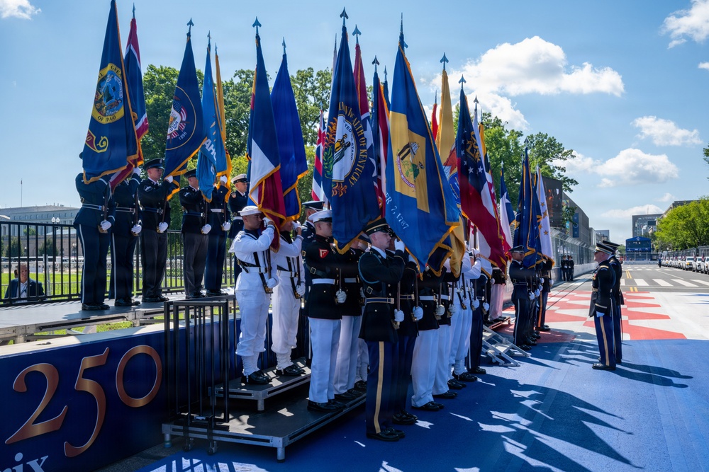 A formal military color guard ceremony is taking place outdoors, featuring personnel from multiple U.S. military branches carrying numerous state and organizational flags. The event appears to be connected to a '250' anniversary commemoration, likely related to the United States' 250th anniversary (America250). The ceremony is held on a decorated parade ground or boulevard with a red, white, and blue themed surface.