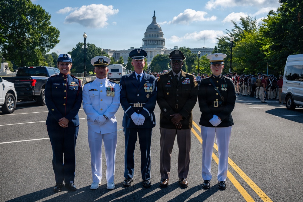 Five U.S. military personnel in dress uniforms representing different branches of the armed forces pose together on a street in Washington, D.C., with the U.S. Capitol Building visible in the background. The individuals appear to be participating in a formal military ceremony or parade event. Historical reenactors in period uniforms are visible in the background to the right.