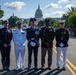 Five U.S. military personnel in dress uniforms representing different branches of the armed forces pose together on a street in Washington, D.C., with the U.S. Capitol Building visible in the background. The individuals appear to be participating in a formal military ceremony or parade event. Historical reenactors in period uniforms are visible in the background to the right.