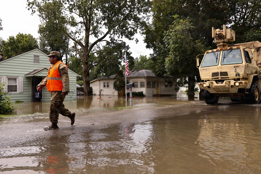 A flood response operation is underway in a residential neighborhood with standing water covering streets and yards. A person in military camouflage uniform and an orange high-visibility vest is wading through floodwater alongside a tan military tactical vehicle. An American flag is visible in the background near partially submerged homes.