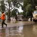 A flood response operation is underway in a residential neighborhood with standing water covering streets and yards. A person in military camouflage uniform and an orange high-visibility vest is wading through floodwater alongside a tan military tactical vehicle. An American flag is visible in the background near partially submerged homes.