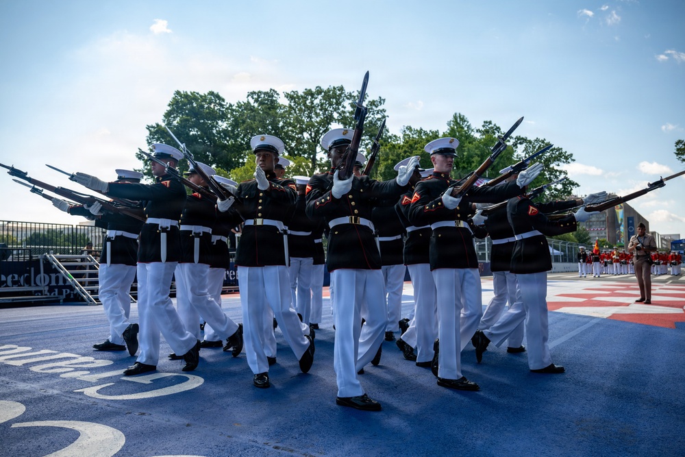 A precision drill team, identifiable as U.S. Marines based on their distinctive dress blue uniforms with white covers and white trousers, performs a ceremonial rifle drill routine. The Marines are executing a synchronized movement with bayonet-tipped rifles extended outward in a diagonal formation. The event appears to take place on a decorated outdoor performance surface, likely during a public ceremonial event.