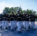 A precision drill team, identifiable as U.S. Marines based on their distinctive dress blue uniforms with white covers and white trousers, performs a ceremonial rifle drill routine. The Marines are executing a synchronized movement with bayonet-tipped rifles extended outward in a diagonal formation. The event appears to take place on a decorated outdoor performance surface, likely during a public ceremonial event.