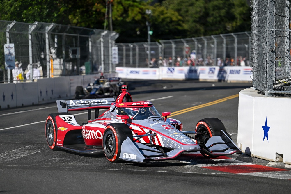 An IndyCar race is taking place on a street circuit, with a prominently featured red and silver Astemo-sponsored Chevrolet IndyCar navigating a tight corner near concrete barriers. The car displays American flag livery elements and carries the 'New Garden' driver name on the cockpit surround. Additional race cars and spectators are visible in the background along the fenced street circuit.