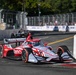 An IndyCar race is taking place on a street circuit, with a prominently featured red and silver Astemo-sponsored Chevrolet IndyCar navigating a tight corner near concrete barriers. The car displays American flag livery elements and carries the 'New Garden' driver name on the cockpit surround. Additional race cars and spectators are visible in the background along the fenced street circuit.
