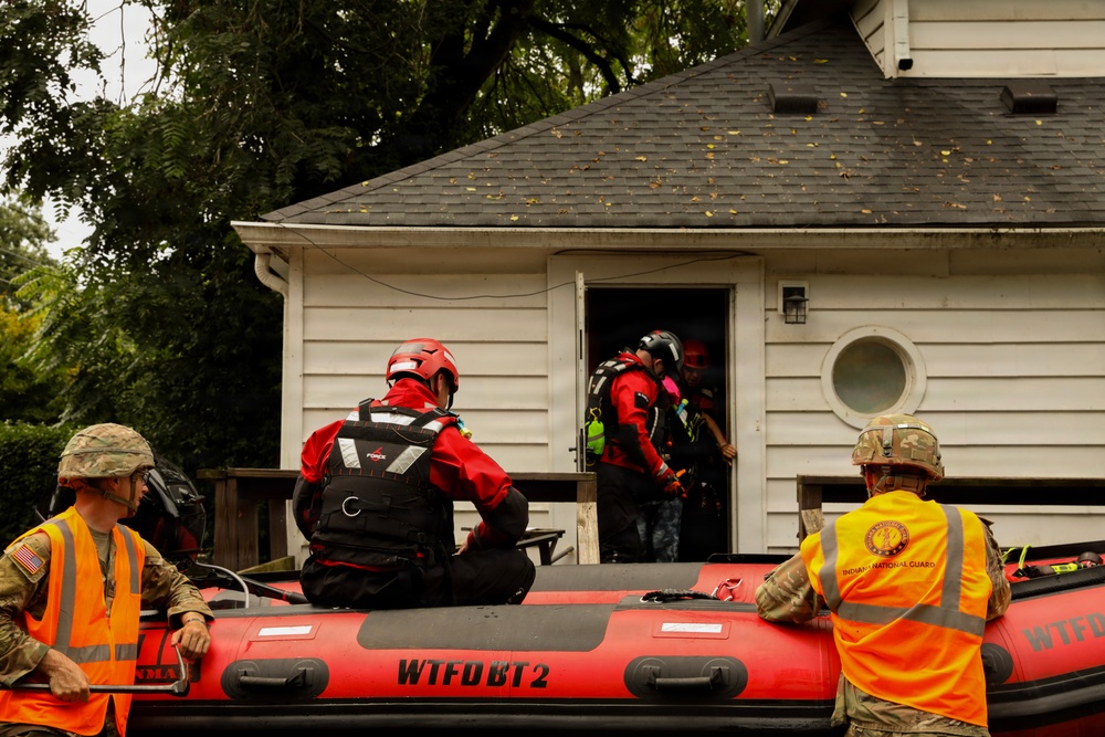 A flood rescue operation is underway, with emergency responders using an inflatable rescue boat marked 'WTFD BT2' to reach a residential structure. Personnel from what appears to be the Indiana National Guard and civilian rescue teams in red gear are working together at the entrance of a white house. Rescuers in red helmets and dry suits are entering or exiting the building, suggesting an active evacuation or welfare check.