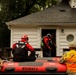 A flood rescue operation is underway, with emergency responders using an inflatable rescue boat marked 'WTFD BT2' to reach a residential structure. Personnel from what appears to be the Indiana National Guard and civilian rescue teams in red gear are working together at the entrance of a white house. Rescuers in red helmets and dry suits are entering or exiting the building, suggesting an active evacuation or welfare check.