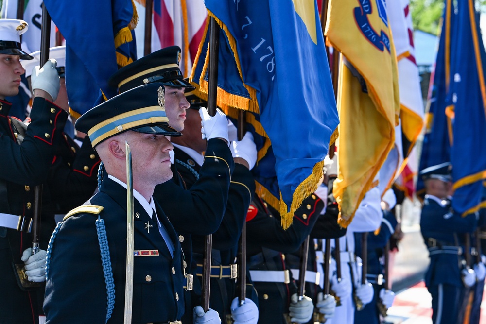 A formal U.S. military joint color guard ceremony is depicted, featuring personnel from multiple branches of the armed forces standing at attention in dress uniforms. Soldiers, Marines, and Air Force members are visible holding ceremonial flags with gold fringe, including what appear to be state and military unit guidons. The scene suggests an official state or military honors ceremony, likely at a prominent government or military installation.