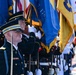 A formal U.S. military joint color guard ceremony is depicted, featuring personnel from multiple branches of the armed forces standing at attention in dress uniforms. Soldiers, Marines, and Air Force members are visible holding ceremonial flags with gold fringe, including what appear to be state and military unit guidons. The scene suggests an official state or military honors ceremony, likely at a prominent government or military installation.